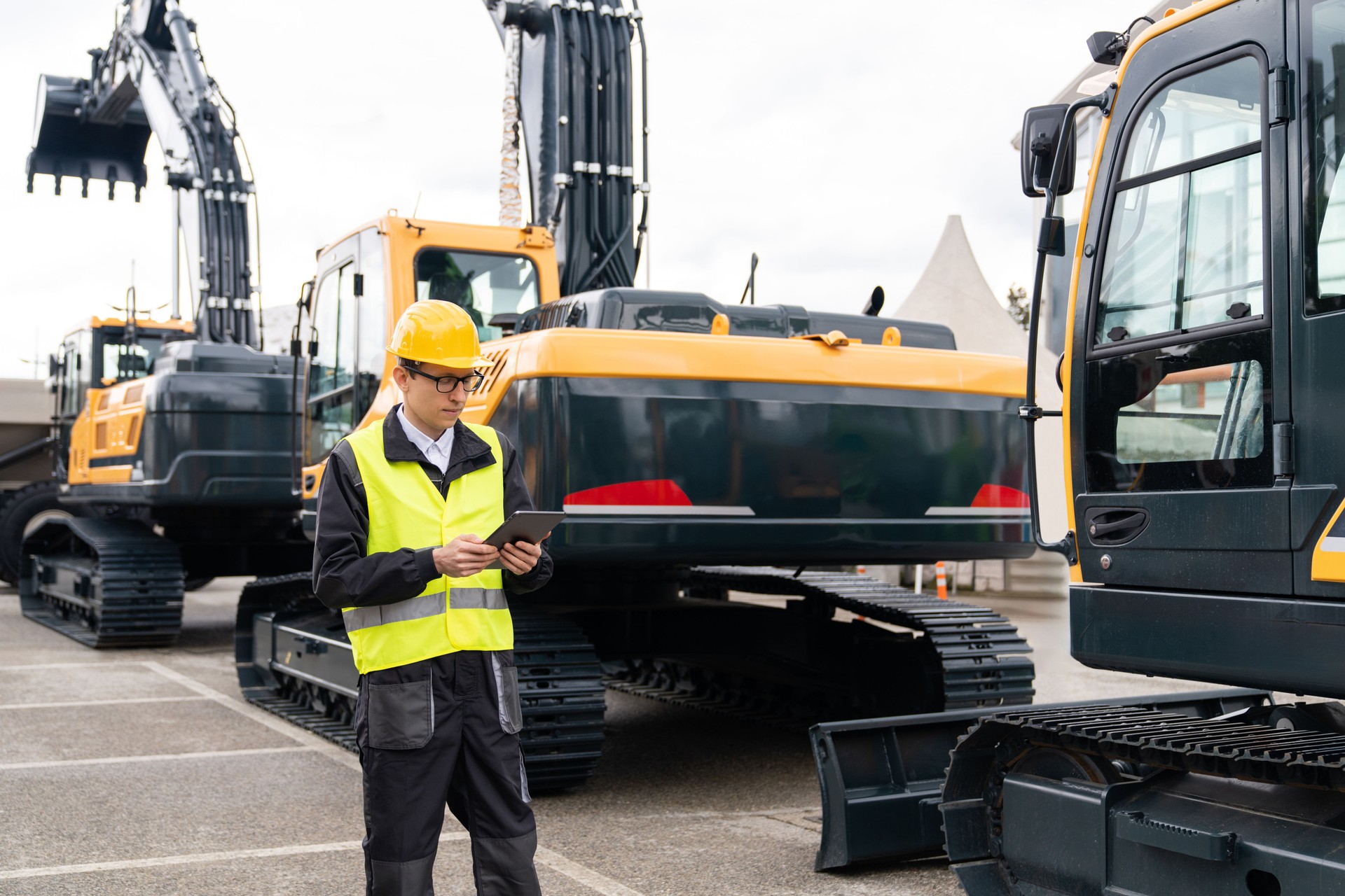 Engineer in a helmet with a digital tablet stands next to construction excavators